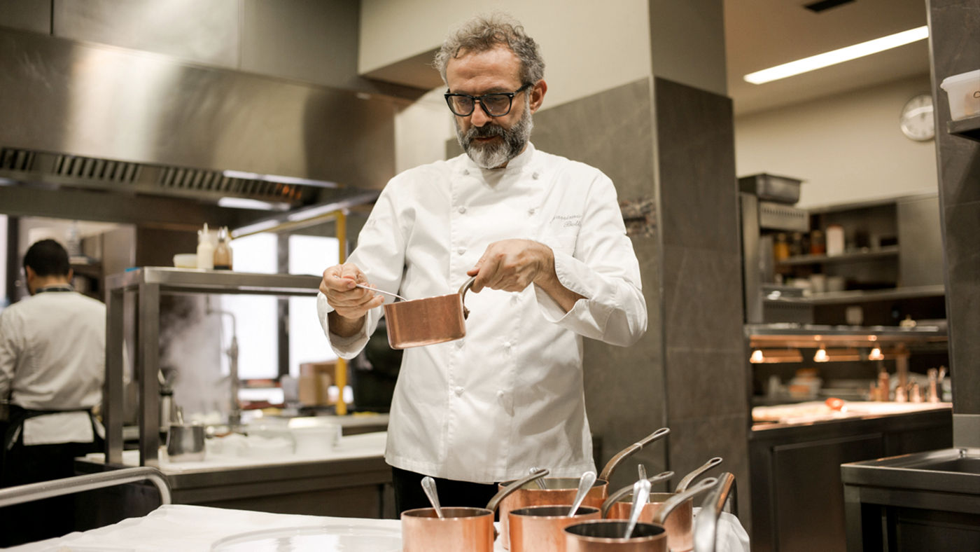 Chef Bottura holding a pot with one hand and a utensil with the other inside a restaurant kitchen