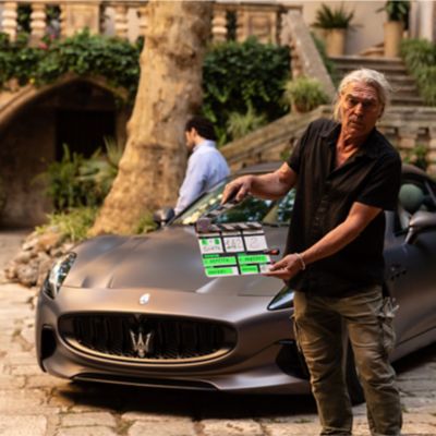 A director on set with a Maserati in Puglia, ready to shoot a scene, with trees and a historic building in the background.