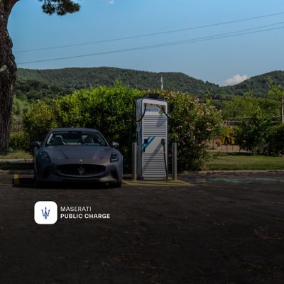 Two Maserati GranTurismo Folgore EVs positioned next to public charging points with a pine tree and hills behind them